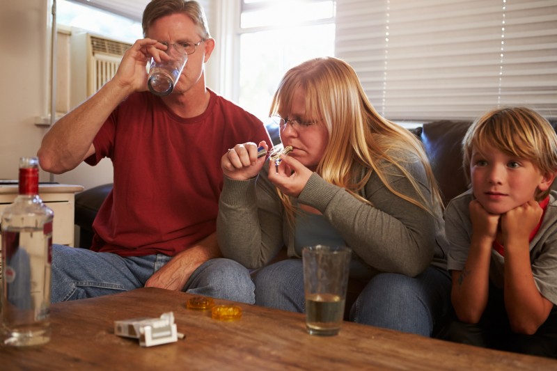 Parents Sit On Sofa With Children Taking Drugs And Drinking